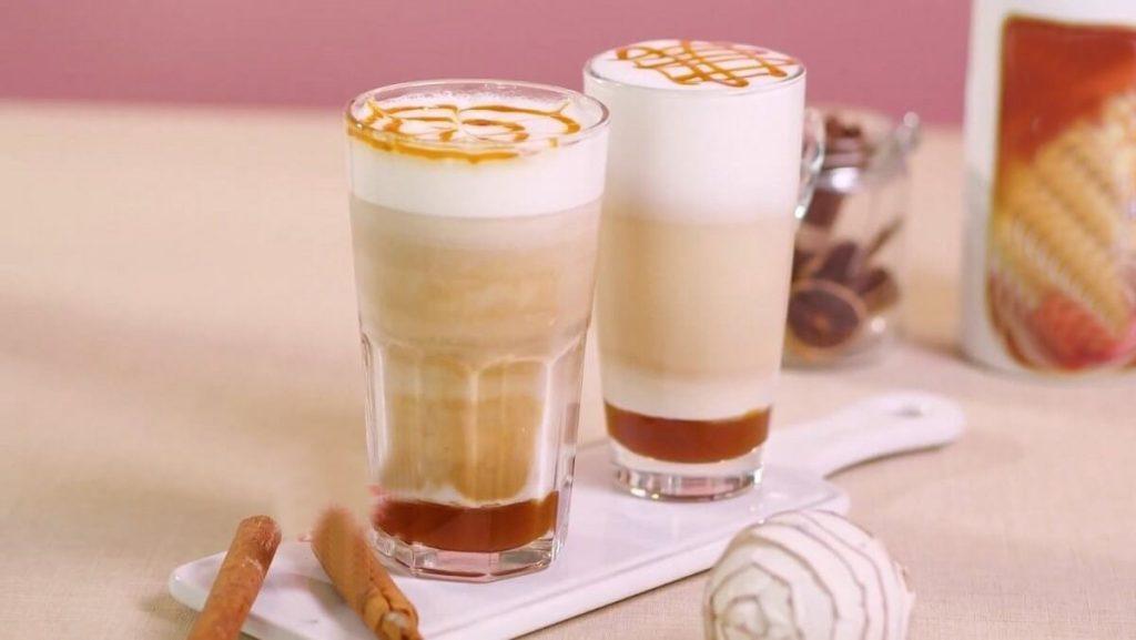 Hai ly cà phê latte với lớp bọt sữa và caramel trên mặt, đặt trên bàn cùng bánh và thanh quế.