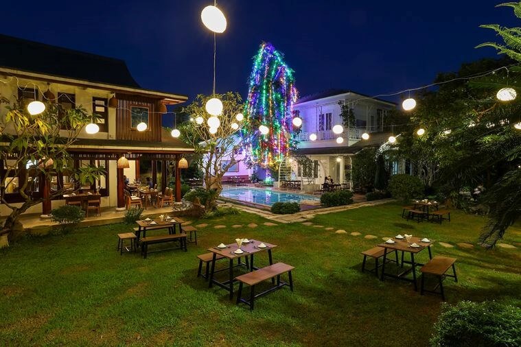 Khu vườn buổi tối được trang trí đèn lấp lánh, bàn ghế gỗ, cây cối xanh tươi, tạo không gian ấm cúng và thư giãn.