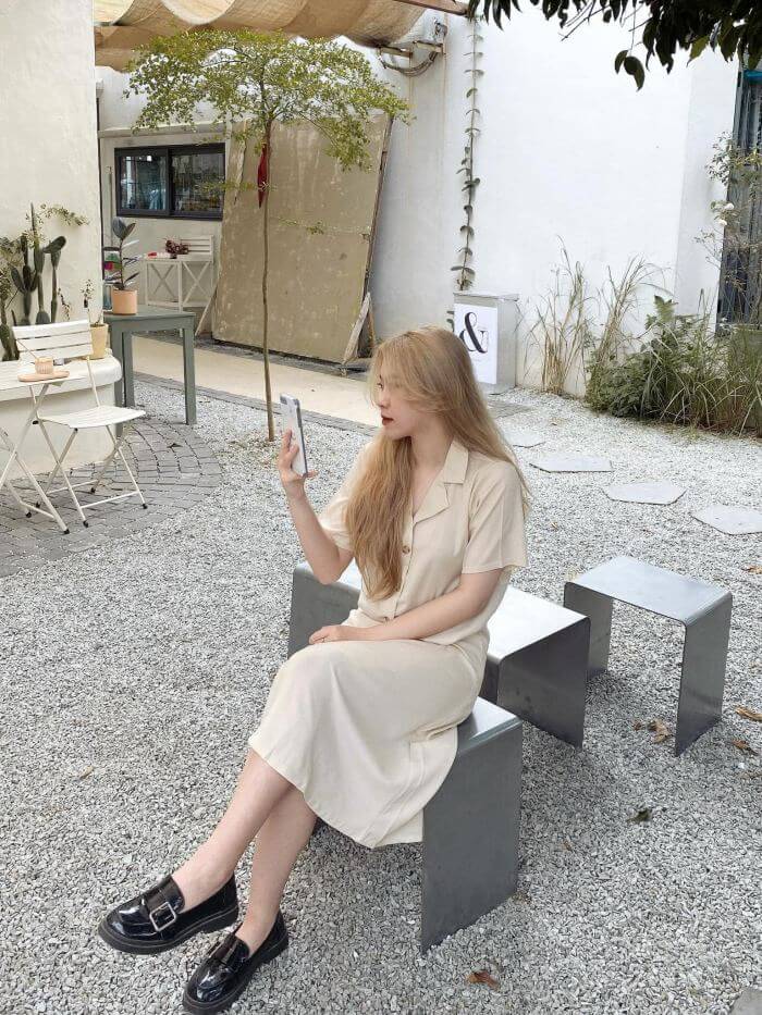 Người phụ nữ ngồi trên ghế kim loại ngoài trời, cầm điện thoại, xung quanh là cây và bàn ghế nhỏ.