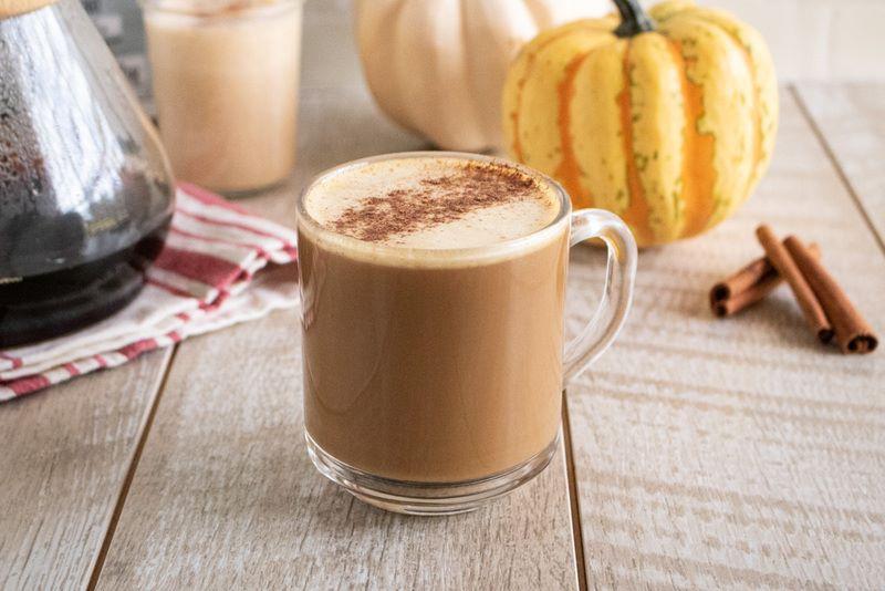 Ly cà phê với bọt sữa và bột quế, xung quanh có bí ngô và thanh quế, trên bàn gỗ.