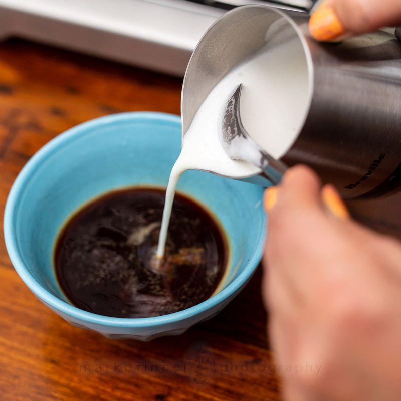 Người đang rót sữa vào cốc đựng cà phê màu xanh trên bàn gỗ.