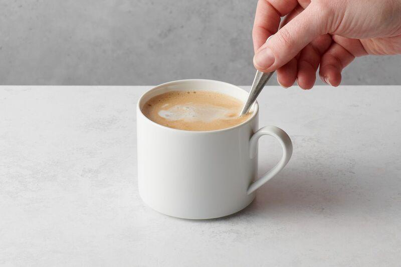 Tay cầm thìa khuấy tách cà phê trên bàn trắng, nền xám.