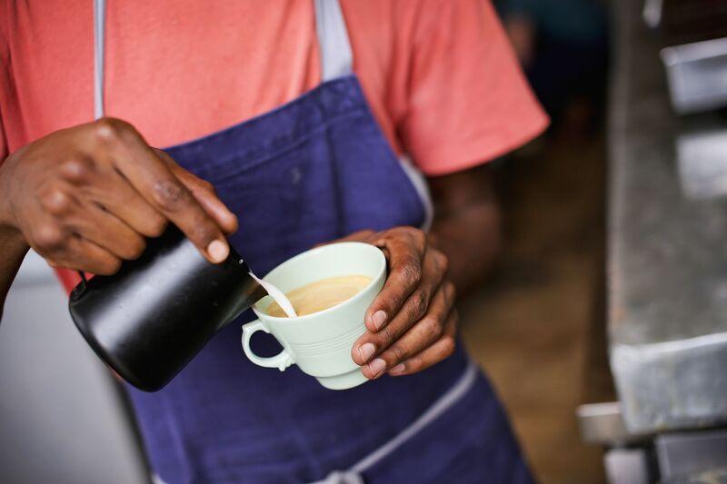 Người mặc tạp dề xanh đang r&oacute;t sữa từ b&igrave;nh đen v&agrave;o t&aacute;ch đồ uống m&agrave;u be.