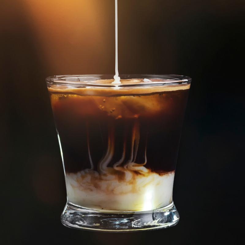 D&ograve;ng sữa trắng đang chảy v&agrave;o ly c&agrave; ph&ecirc; đen, tạo hiệu ứng h&ograve;a quyện độc đ&aacute;o trong &aacute;nh s&aacute;ng mờ ảo.