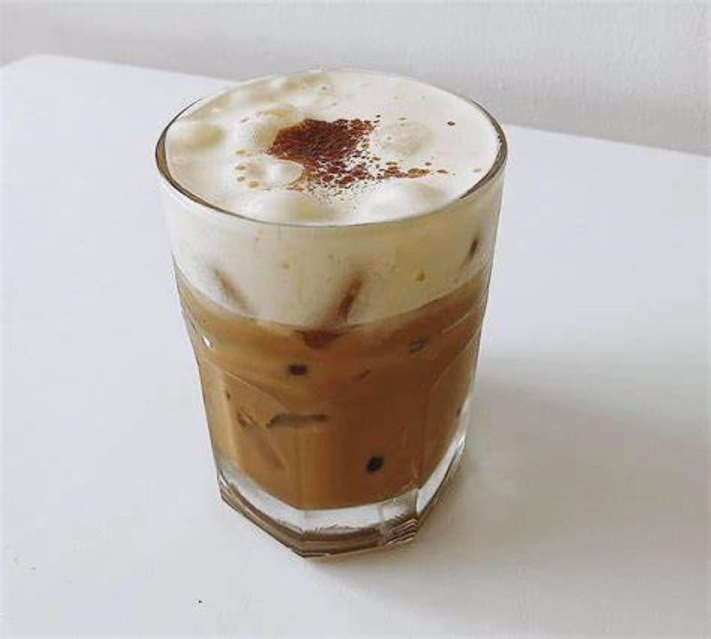 Ly cà phê sữa đá với lớp bọt sữa và chút bột cacao trên bề mặt, đặt trên bàn trắng.