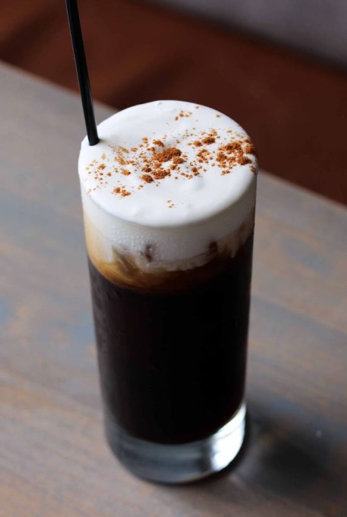 Ly cà phê đen với lớp kem trắng và bột cacao, ống hút đen đặt trên mặt bàn gỗ.