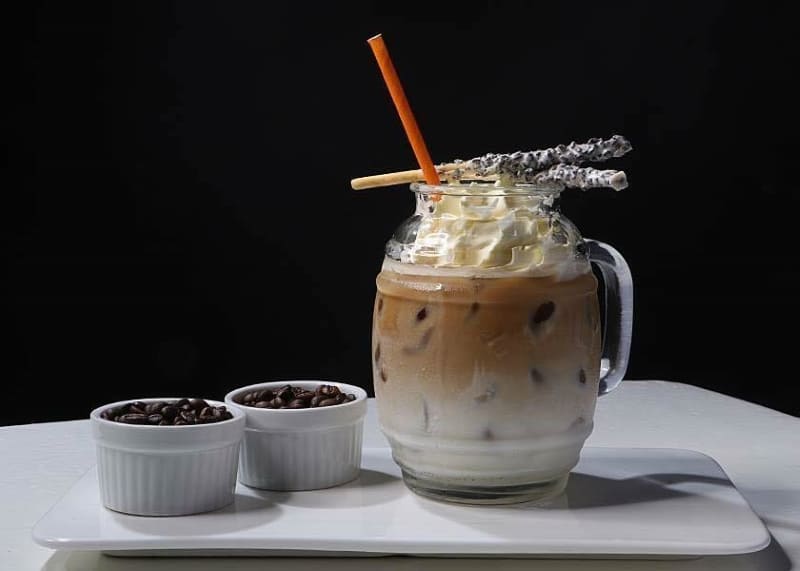 Ly cà phê sữa đá có kem tươi, ống hút cam và bánh que, bên cạnh là hai chén nhỏ đựng hạt cà phê.