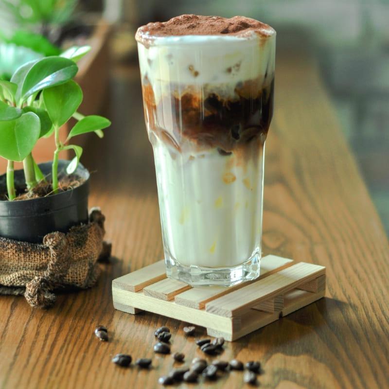 Ly cà phê sữa đá có lớp bọt cacao, bên cạnh là chậu cây nhỏ trên bàn gỗ với vài hạt cà phê rơi vãi.