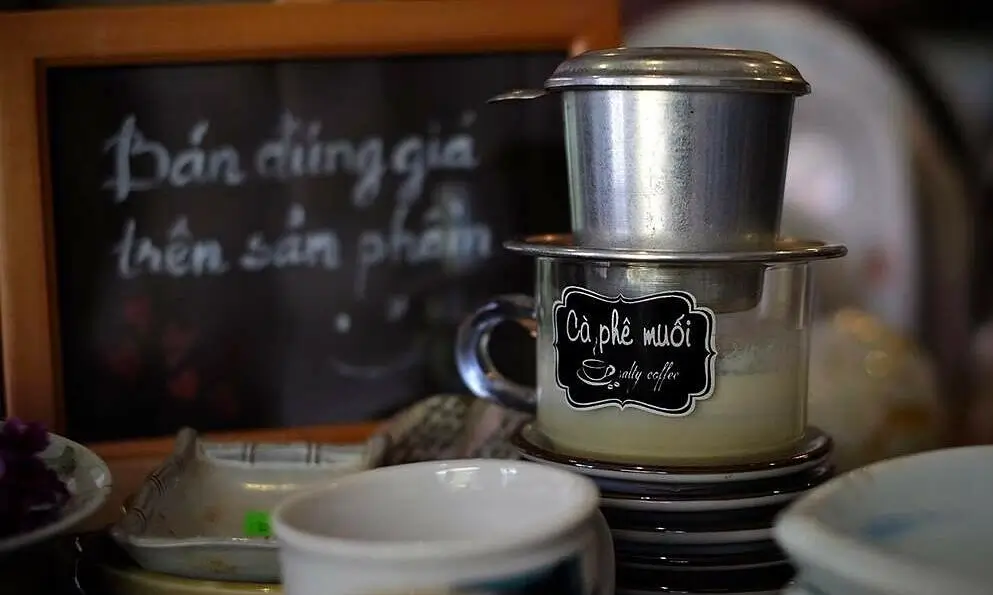 Phin cà phê muối trên bàn, bên cạnh bảng đen ghi "Bán đúng giá trên sản phẩm".