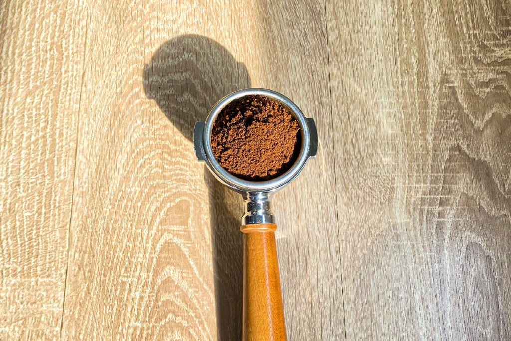 cach-lam-cappuccino Tay cầm pha cà phê chứa bột cà phê tươi nằm trên mặt bàn gỗ, ánh sáng chiếu tạo bóng.