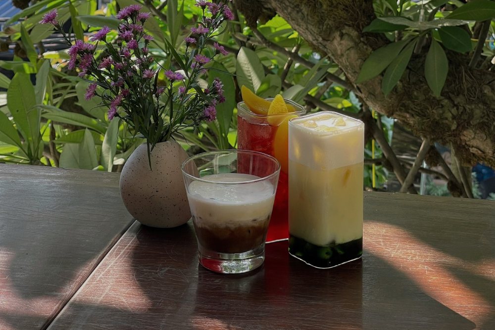 Khám Phá 11 Quán Cafe Đội Cấn Tuyệt Vời Cho Thư Giãn Và Làm Việc 31 Ba ly nước uống đa dạng trên bàn gỗ, bên cạnh là bình hoa nhỏ; nền là cây xanh.