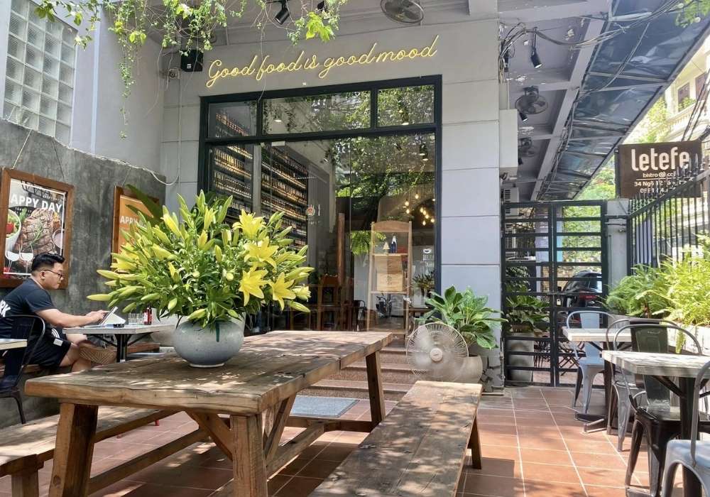 Khám Phá Top 10 Quán Cafe Láng Hạ: View Đẹp, Menu Hấp Dẫn, Giá Cực Hợp Lý 17 Quán cà phê ngoài trời với bàn gỗ, cây xanh và bảng hiệu. Một người ngồi làm việc trên máy tính.