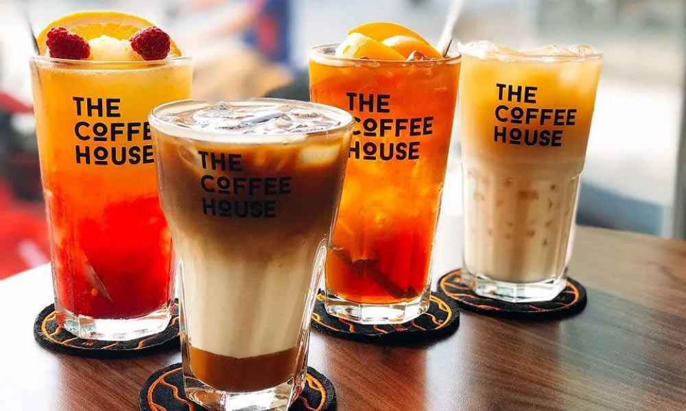 Khám Phá 14 Quán Cafe Hồ Tùng Mậu: Điểm Chill Lý Tưởng Tại Cầu Giấy 4 Bốn ly đồ uống đa dạng màu sắc từ The Coffee House, đặt trên bàn gỗ, mỗi ly có logo thương hiệu in trên.