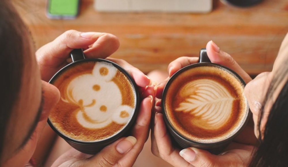 co-gai-uong-ca-phe-cappuccino Hai người cầm cốc cà phê với hình vẽ nghệ thuật, một hình gấu và một hình lá.