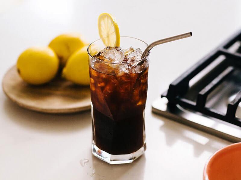 Ly c&agrave; ph&ecirc; đ&aacute; với l&aacute;t chanh tr&ecirc;n miệng ly, ống h&uacute;t kim loại, b&ecirc;n cạnh l&agrave; đĩa chanh v&agrave;ng v&agrave; bếp ga.