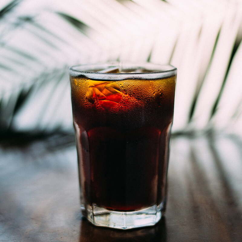 Ly nước c&oacute; m&agrave;u sẫm với đ&aacute; vi&ecirc;n, đặt tr&ecirc;n b&agrave;n gỗ, nền l&agrave; b&oacute;ng c&acirc;y mờ ảo.