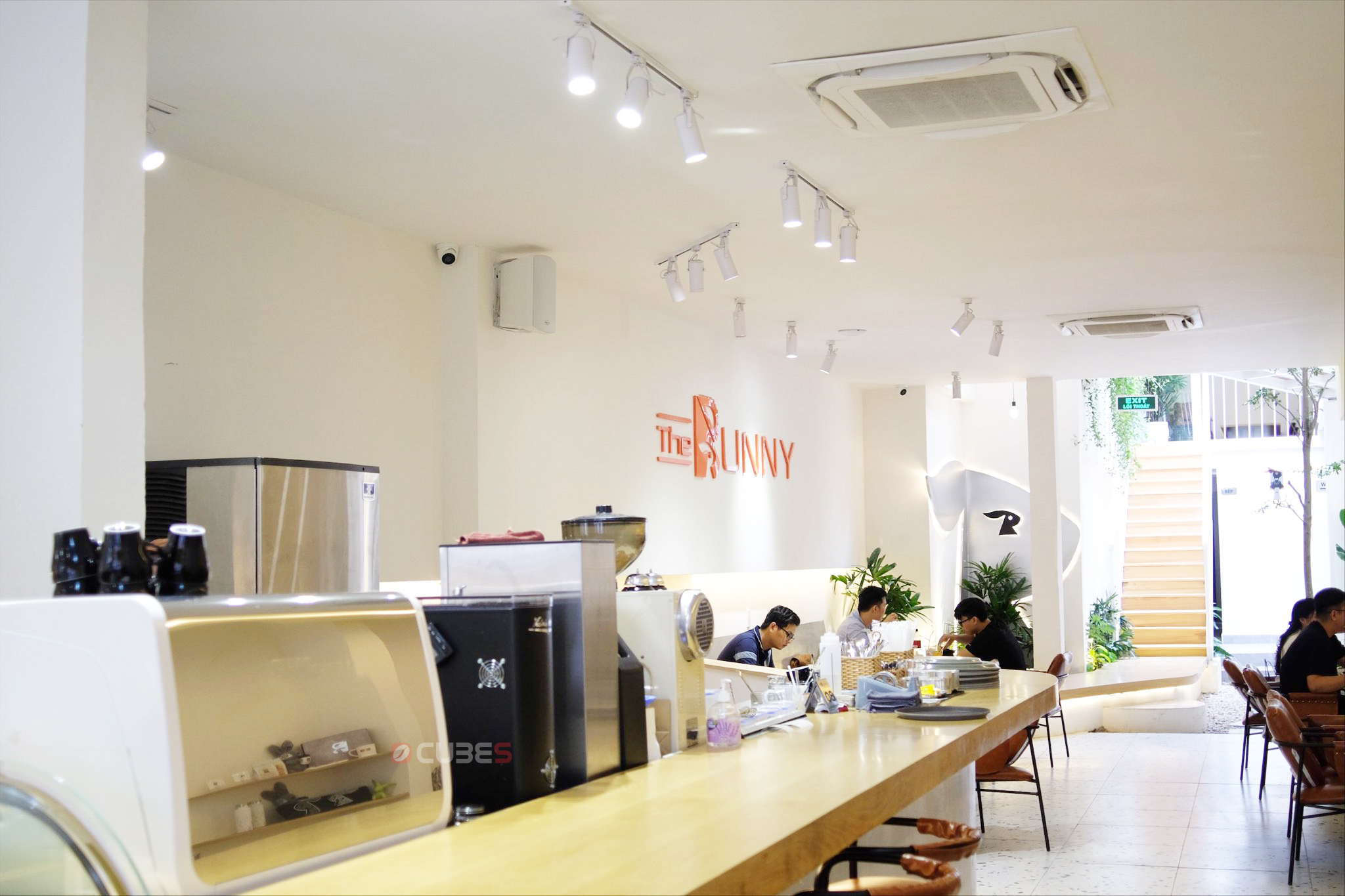 the-bunny-coffee-3-su-dung-may-pha-ca-phe-appia-life-two-group Quán cà phê với bàn dài, vài người ngồi trò chuyện, ánh sáng tự nhiên và cây xanh trang trí.