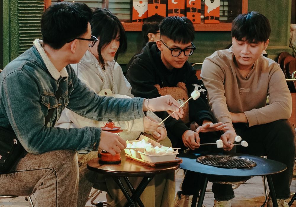 Khám Phá Top 10 Quán Cafe Láng Hạ: View Đẹp, Menu Hấp Dẫn, Giá Cực Hợp Lý 20 Nhóm người ngồi quanh bàn, nướng kẹo marshmallow trên bếp nướng trong không gian ấm cúng.