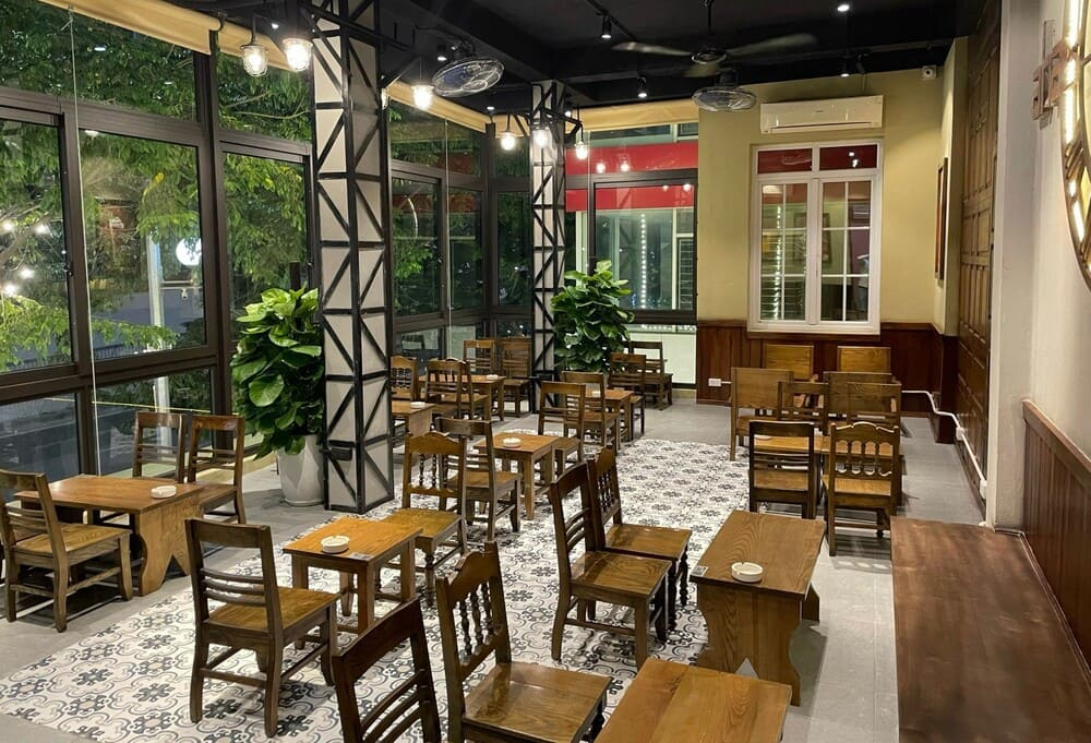 Khám Phá 15 Quán Cafe Hấp Dẫn Trên Đường Nguyễn Thị Định 16 Quán cà phê với bàn ghế gỗ, cửa kính lớn, cây xanh trang trí, không gian thoáng đãng và ánh sáng dịu nhẹ.