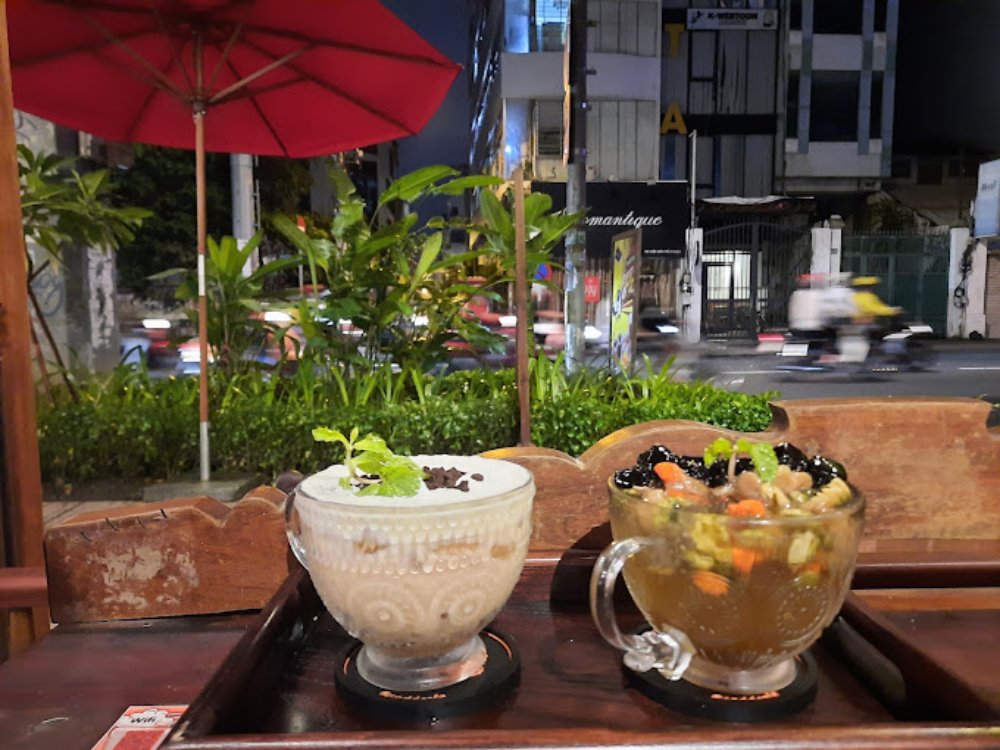 Khám Phá Top 6 Quán Cafe Mở Cửa 24h Tại Quận 3 Cho Người Yêu Thích Cú Đêm 14 Hai ly đồ uống trên bàn gỗ, một ly có cây bạc hà, bên ngoài là khung cảnh đường phố buổi tối.
