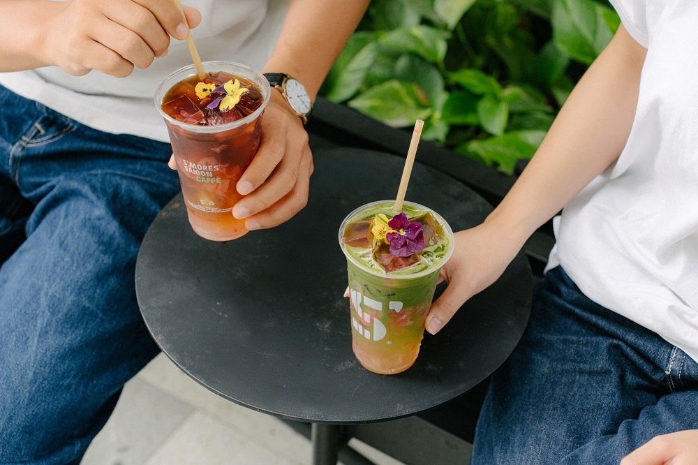 Khám Phá Những Quán Cafe Độc Lạ Thu Hút Giới Trẻ Tại Sài Gòn 15 Hai người cầm ly nước có ống hút và hoa trang trí, ngồi bên bàn tròn màu đen.
