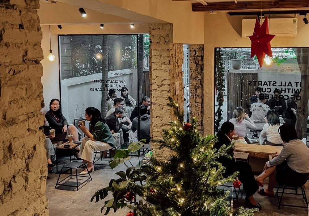 Khám Phá Top Quán Cafe Đẹp Ở Liễu Giai: Đồ Uống Ngon, Không Gian Thư Giãn 16 Quán cà phê ấm cúng với cây thông trang trí, mọi người ngồi trò chuyện bên trong và ngoài cửa kính lớn.