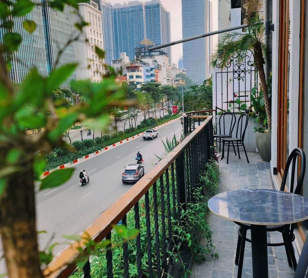 Khám Phá Top Quán Cafe Đẹp Ở Liễu Giai: Đồ Uống Ngon, Không Gian Thư Giãn 14 Ban công nhìn ra đường phố, với xe cộ di chuyển, cây xanh và tòa nhà cao tầng phía xa.