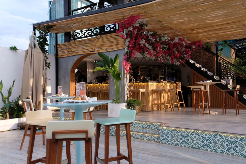 Khám Phá Top 10 Quán Rooftop Quận 1: View Đẹp Ngắm Thành Phố, Trải Nghiệm Thư Giãn Tuyệt Đỉnh 8 Quán cà phê ngoài trời với bàn ghế gỗ, trang trí hoa giấy và quầy bar bên trong, không gian thoáng đãng.