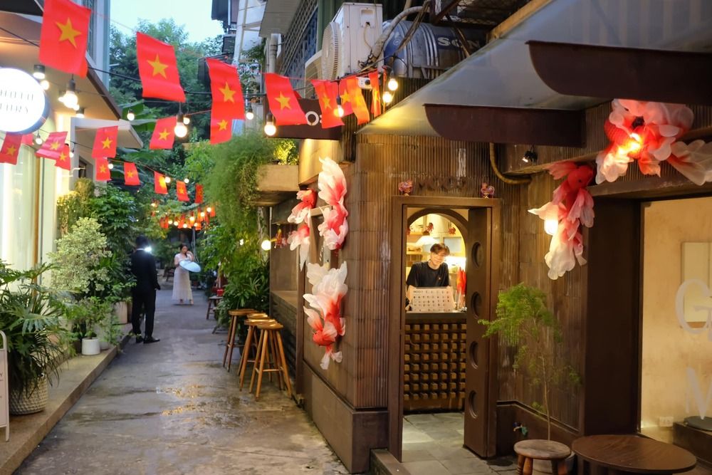 Khám Phá 10+ Quán Cafe Đẹp Ở Đặng Văn Ngữ: Đồ Uống Ngon, Giá Cả Hấp Dẫn 21 Con hẻm nhỏ trang trí cờ đỏ sao vàng, đèn lấp lánh, và hoa giấy, có người đứng bên quầy hàng.