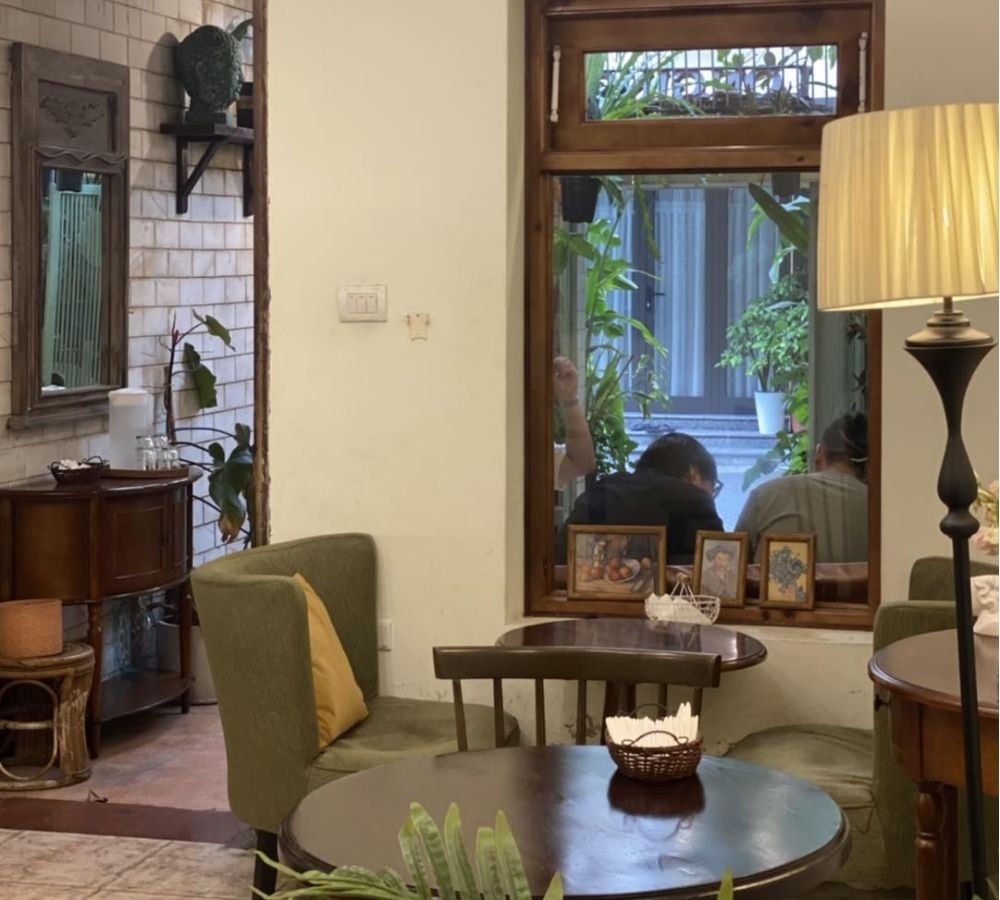 Khám Phá Top Quán Cafe Đẹp Ở Liễu Giai: Đồ Uống Ngon, Không Gian Thư Giãn 21 Góc quán cà phê ấm cúng với bàn ghế gỗ, đèn bàn và cửa sổ nhìn ra khu vườn xanh mát bên ngoài.