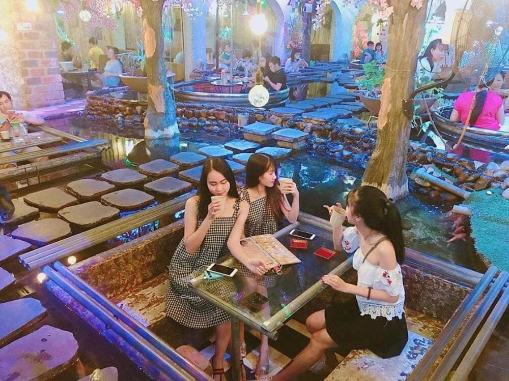 Ba người ngồi tại bàn trong quán cà phê có không gian trang trí bằng cây xanh và đèn trang trí.