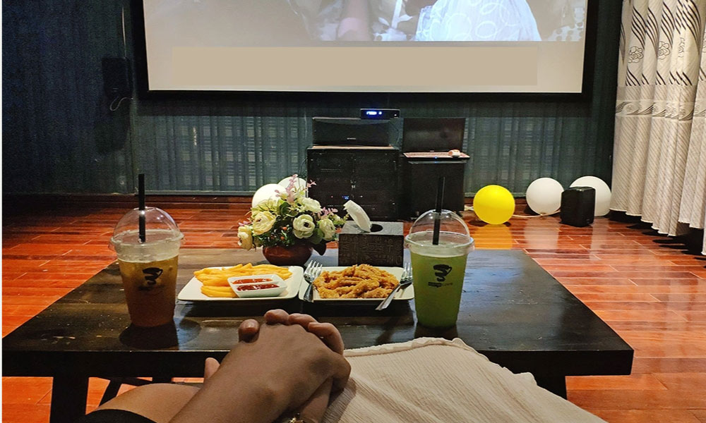 Khám Phá 14 Quán Cafe Hồ Tùng Mậu: Điểm Chill Lý Tưởng Tại Cầu Giấy 20 Hai ly nước và đồ ăn nhẹ trên bàn trước màn hình chiếu phim, trang trí với hoa và bóng bay trong phòng.