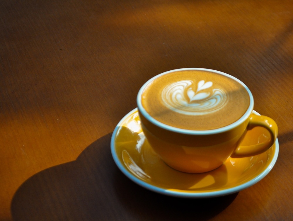 15 Quán Cafe Chùa Láng Với Không Gian Thư Giãn Tuyệt Đẹp 15 Tách cà phê với họa tiết latte art trong chiếc cốc màu vàng, đặt trên bàn gỗ dưới ánh sáng tự nhiên.