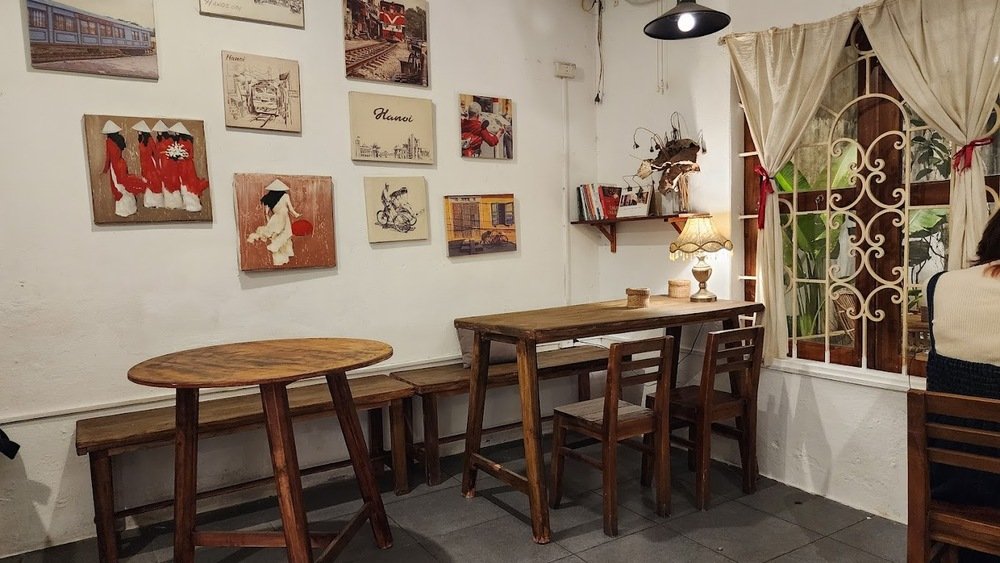Khám Phá 10+ Quán Cafe Đẹp Ở Đặng Văn Ngữ: Đồ Uống Ngon, Giá Cả Hấp Dẫn 10 Quán cà phê với bàn ghế gỗ, tranh treo tường và cửa sổ có rèm, tạo không gian ấm cúng và thư giãn.