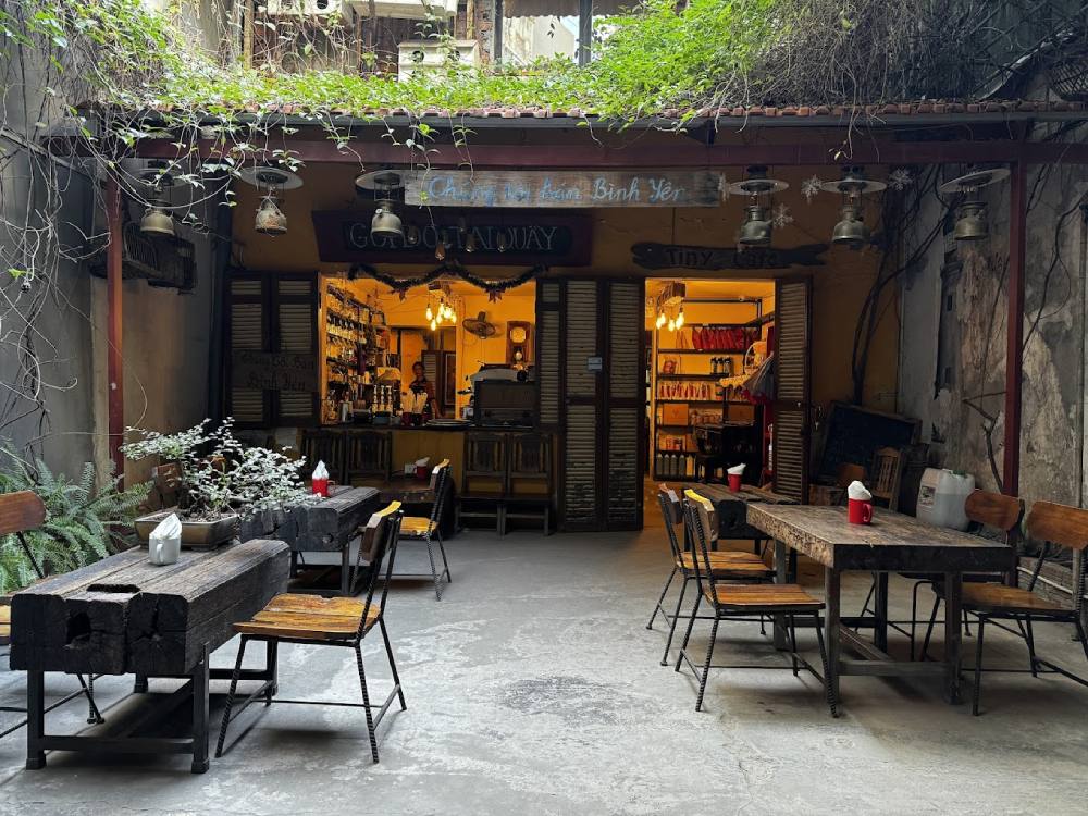 Khám Phá 11 Quán Cafe Đội Cấn Tuyệt Vời Cho Thư Giãn Và Làm Việc 4 Quán cà phê nhỏ với bàn ghế gỗ, cây xanh, và ánh sáng ấm áp từ bên trong, tạo không gian ấm cúng.