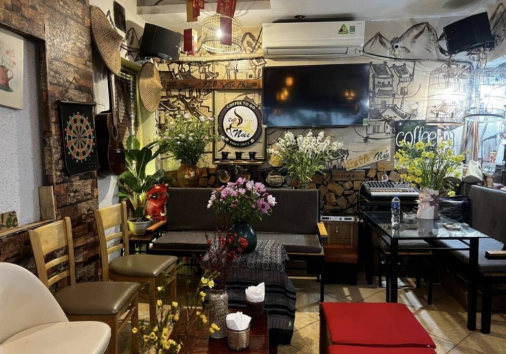 Khám Phá Top Quán Cafe Đẹp Ở Liễu Giai: Đồ Uống Ngon, Không Gian Thư Giãn 19 Quán cà phê với nội thất ấm cúng, trang trí bằng hoa, nhạc cụ và tranh tường. Có bảng phi tiêu và bộ ghế ngồi.