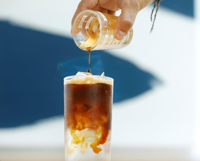 Một người đang rót cà phê từ cốc đong vào ly đá, tạo nên lớp màu nâu hòa quyện.