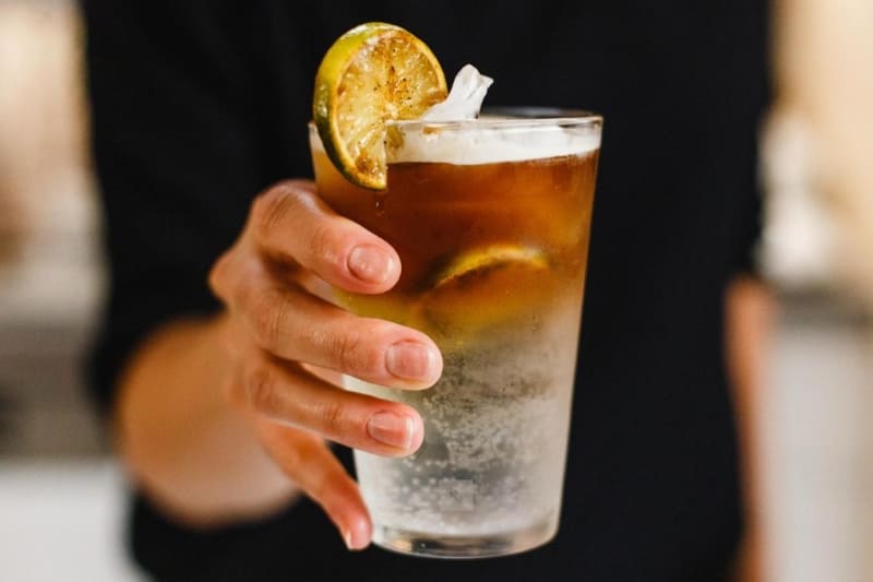 Tay cầm ly đồ uống có chanh cắt lát và đá, nền mờ.