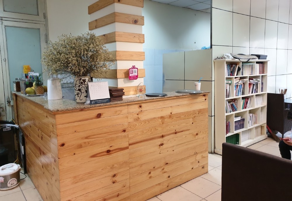 Khám Phá 11 Quán Cafe Sách Độc Đáo Tại Cầu Giấy Hút Hồn Độc Giả 6 Quầy gỗ trong phòng với kệ sách và cây trang trí, cạnh một người đang ngồi đọc sách.