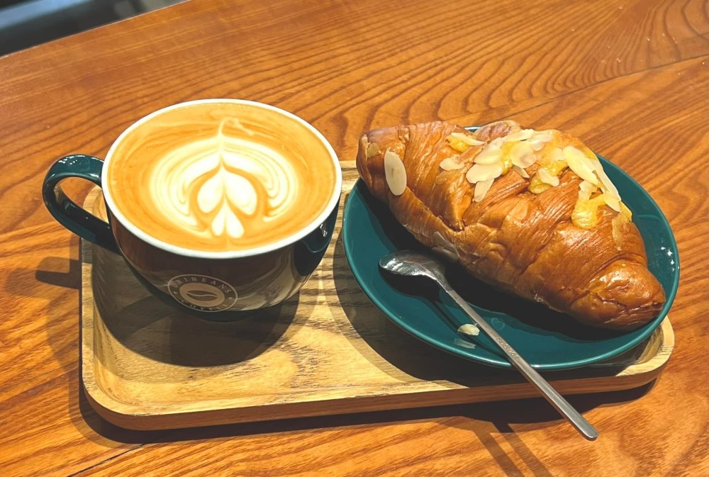 Khám Phá Top 10 Quán Cafe Giảng Võ Hấp Dẫn Nhất Tại Hà Nội 20 Tách cà phê nghệ thuật và bánh sừng bò hạnh nhân trên đĩa xanh, đặt trên khay gỗ.