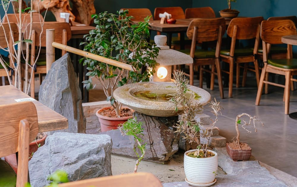 Khám Phá 15+ Quán Cafe View Đẹp, Đồ Uống Ngon Tại Hoàng Cầu 23 Một góc quán cà phê với bàn ghế gỗ, trang trí bằng cây xanh và tiểu cảnh nước phong cách Nhật Bản.