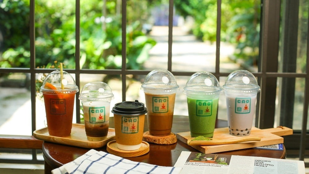 Khám Phá 15+ Quán Cafe View Đẹp, Đồ Uống Ngon Tại Hoàng Cầu 9 Năm ly đồ uống đa dạng trên bàn gỗ, bên cửa sổ sáng, có cây xanh bên ngoài.