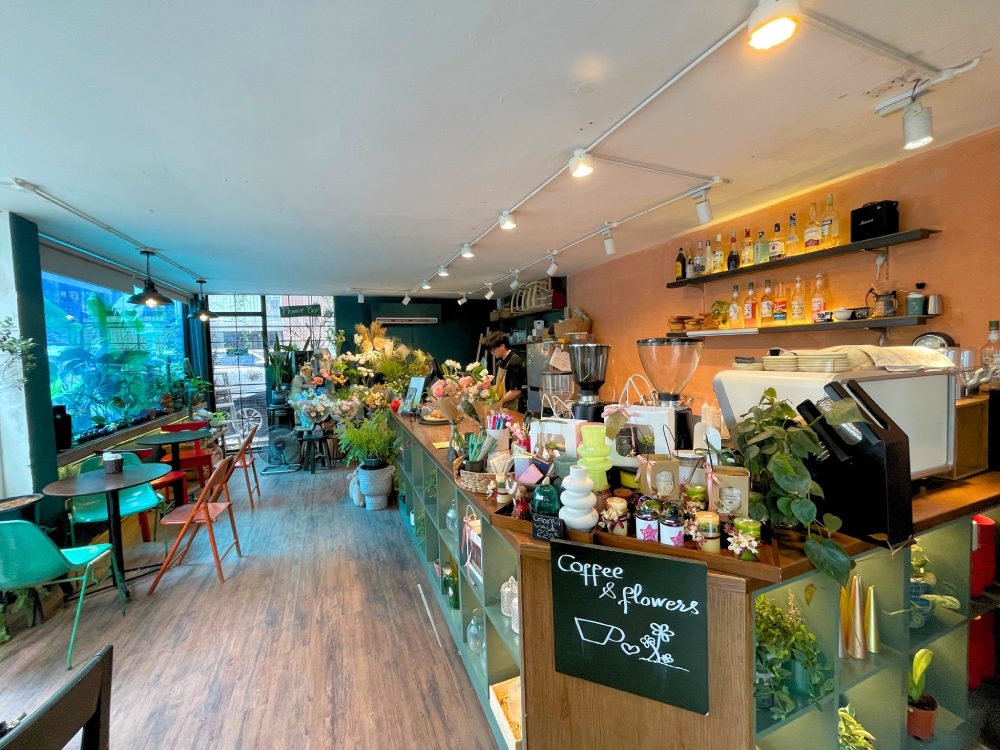 Khám Phá 11 Quán Cafe Đội Cấn Tuyệt Vời Cho Thư Giãn Và Làm Việc 27 Quán cà phê với không gian nội thất gỗ, trang trí nhiều cây xanh và hoa, có quầy phục vụ đồ uống và kệ trưng bày.