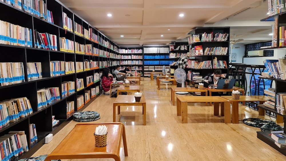 Khám Phá 11 Quán Cafe Sách Độc Đáo Tại Cầu Giấy Hút Hồn Độc Giả 5 Thư viện với kệ sách lớn và bàn ghế gỗ thấp; nhiều người ngồi đọc sách và làm việc trên máy tính.