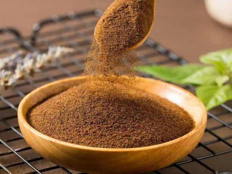 Bột m&agrave;u n&acirc;u được đổ từ muỗng v&agrave;o b&aacute;t gỗ, đặt tr&ecirc;n bề mặt lưới, với l&aacute; c&acirc;y xanh ở nền.