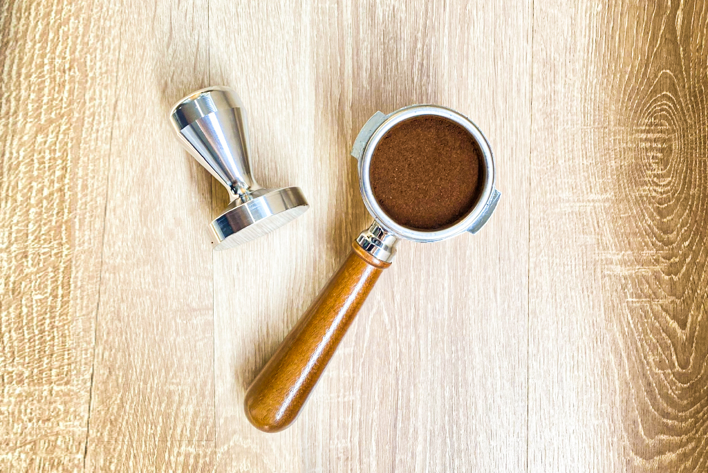 cach-lam-cappuccino Dụng cụ nén cà phê và tay cầm chứa bột cà phê nằm trên bề mặt gỗ sáng màu.