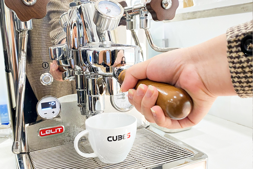 Tay người đang sử dụng m&aacute;y pha c&agrave; ph&ecirc;, với một t&aacute;ch trắng đặt ph&iacute;a dưới v&ograve;i chiết xuất.