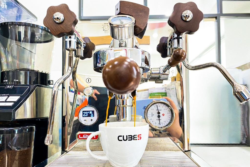 M&aacute;y pha c&agrave; ph&ecirc; đang chảy c&agrave; ph&ecirc; v&agrave;o t&aacute;ch, xung quanh l&agrave; c&aacute;c n&uacute;t điều khiển v&agrave; m&aacute;y đo &aacute;p suất.