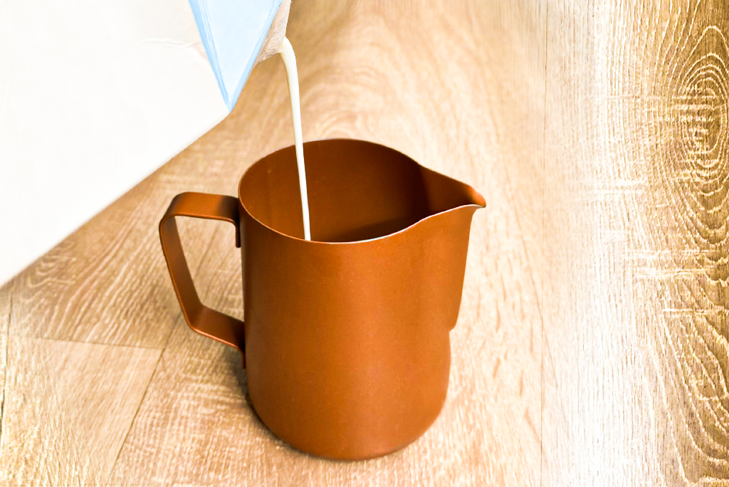 Hộp giấy đang r&oacute;t chất lỏng m&agrave;u trắng v&agrave;o b&igrave;nh kim loại m&agrave;u n&acirc;u tr&ecirc;n nền gỗ.