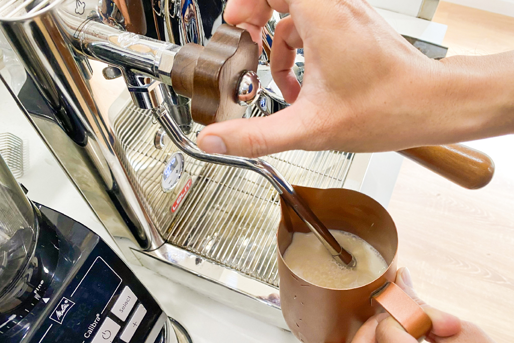 Người đang d&ugrave;ng m&aacute;y pha c&agrave; ph&ecirc; để đ&aacute;nh sữa trong một ca kim loại m&agrave;u n&acirc;u.